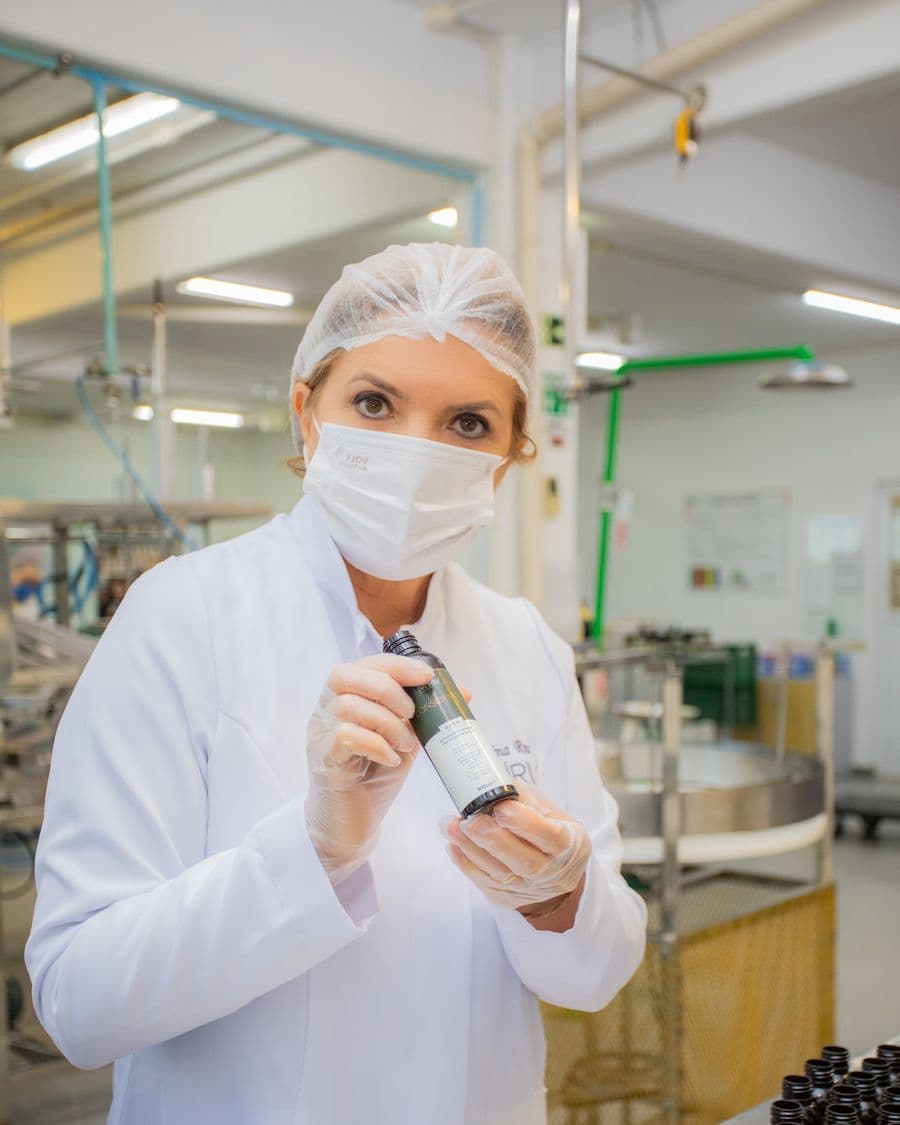 Sabrina Rosa de jaleco e touca no laboratório da fábrica, segurando um frasco